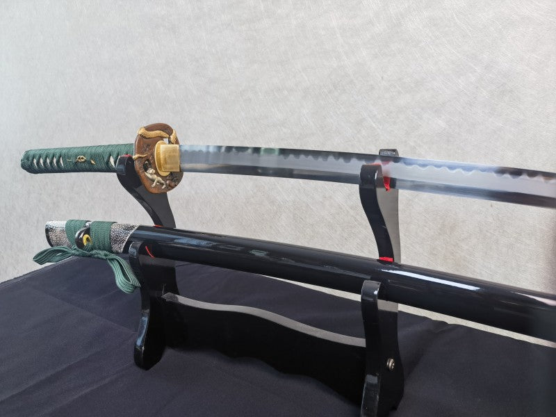 A close-up of the tsuka handle, showing the green silk ito wrap and the brass serpent theme tsuba.