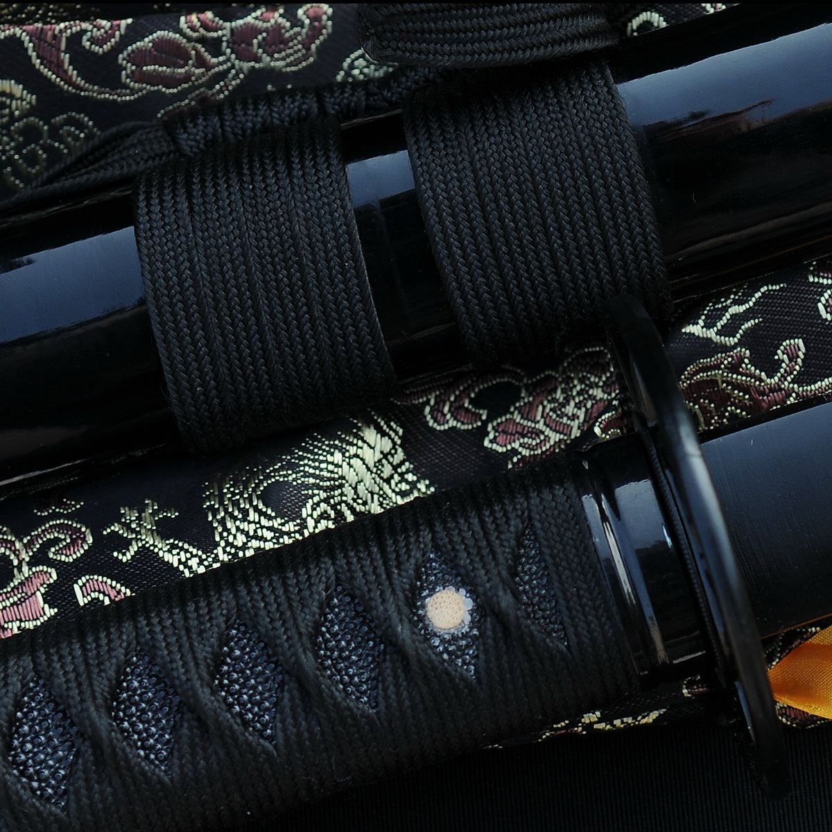 A close-up of the Kuro Akane Katana's handle, showing the black silk ito over black genuine rayskin, alongside the gloss-black saya and brocade sageo.