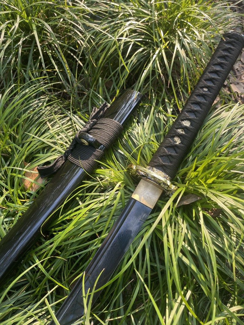 The unsheathed Obsidian Edge Katana, showcasing its full through-hardened, blackened folded steel blade in grass.
