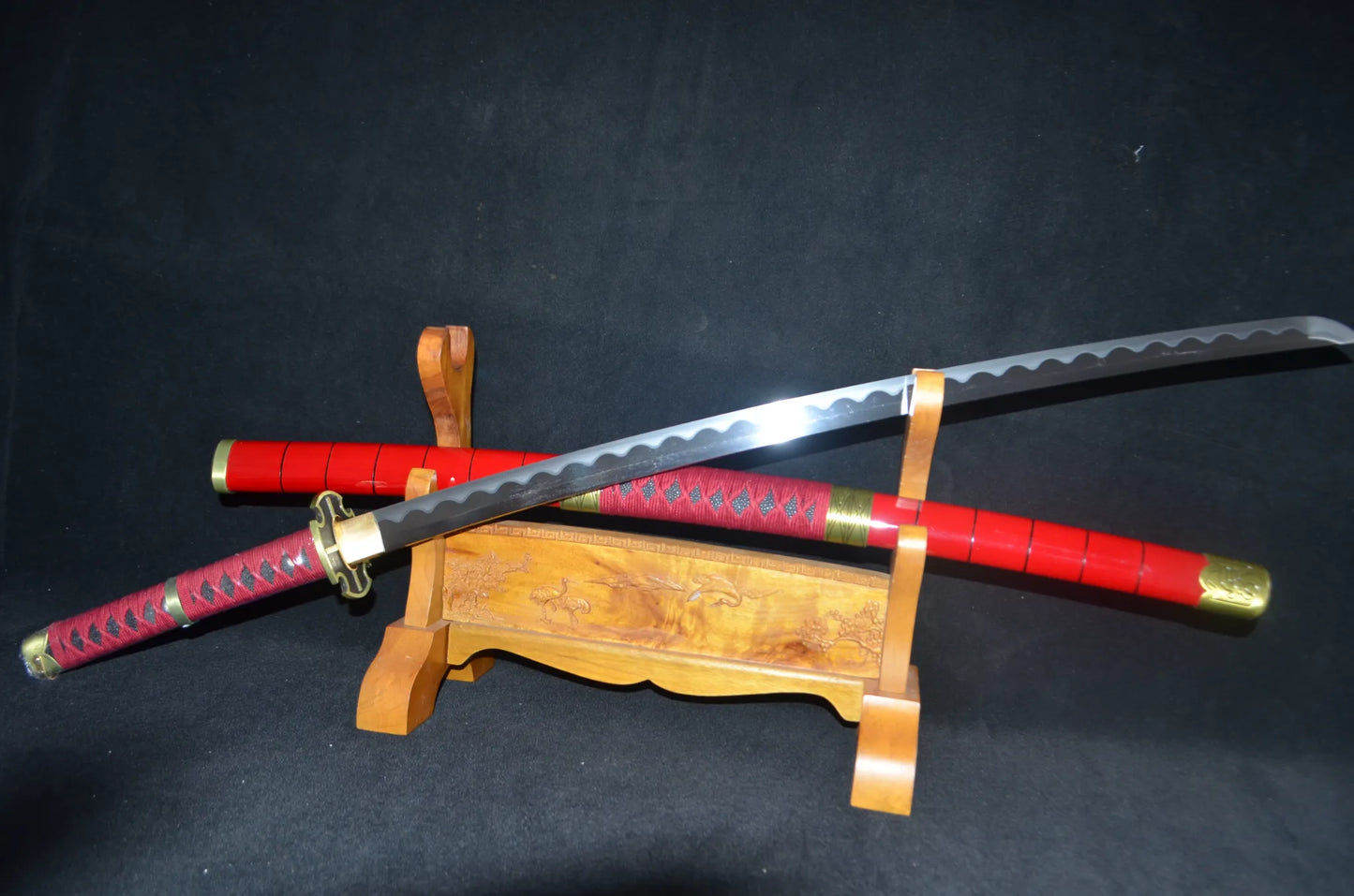 The Sandai Kitetsu katana drawn at an angle, showing the wavy clay-tempered hamon on its polished blade above the red saya and wooden stand.
