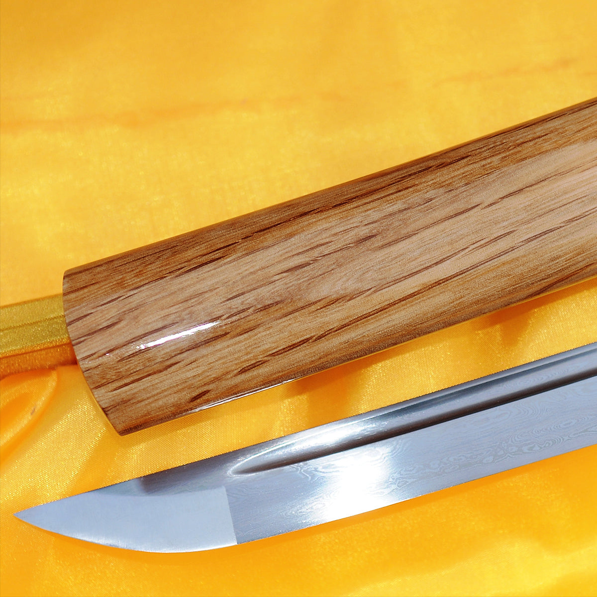 A close-up of the Taka's Sky Katana's folded steel kissaki (tip), lying next to the polished natural hardwood saya which shows its wood grain.
