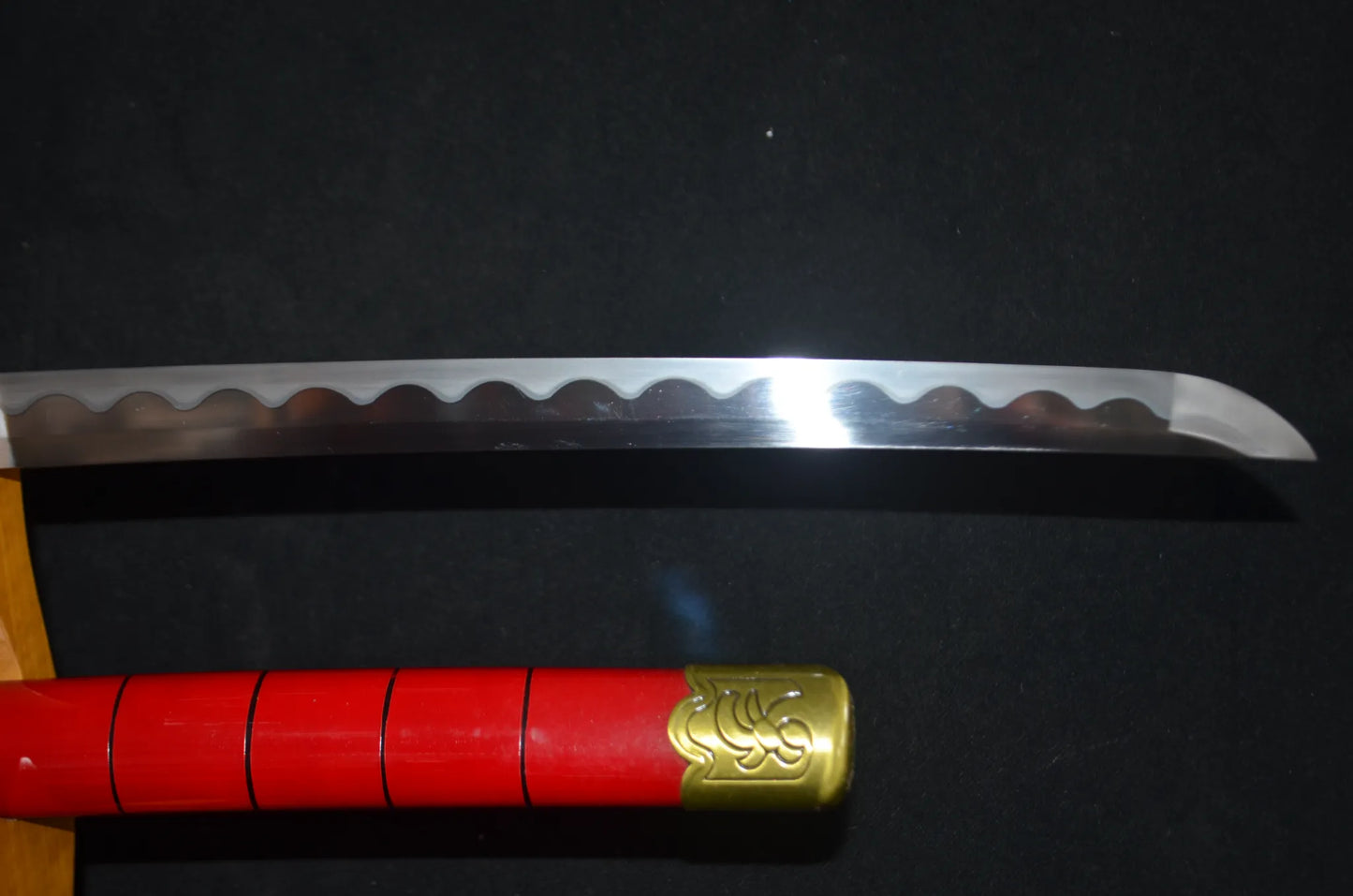 Detail of the Sandai Kitetsu katana's tip and the gold kojiri end-cap on its red saya, with the hamon visible along the blade.
