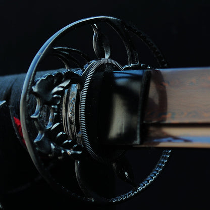 A top-down view of the Kuro Akane Katana's iron sukashi tsuba, showing the botanical vine theme against the red and black folded steel blade.