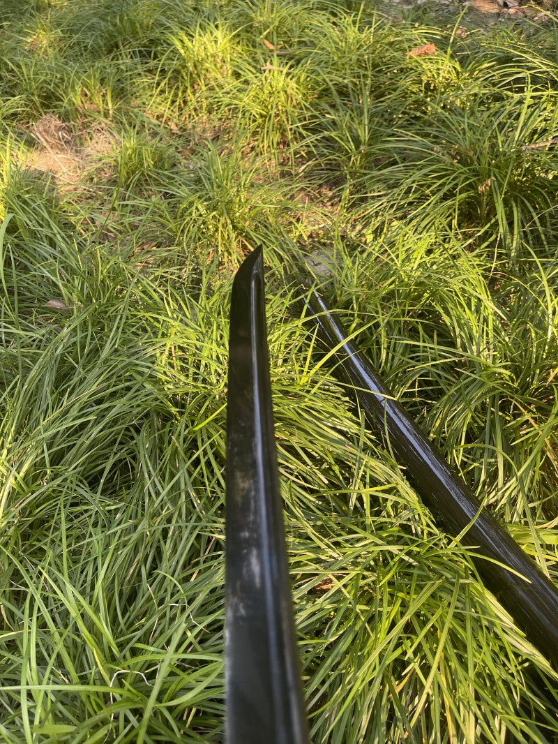 The complete Obsidian Edge Katana, fully sheathed in its gloss-black piano lacquer saya, resting in a grassy setting.