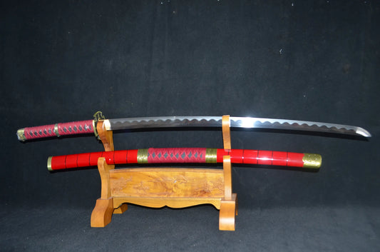 The Sandai Kitetsu "One Piece" katana on a carved wooden stand — 1095 steel blade with a wavy clay-tempered hamon above its glossy red saya with gold fittings.