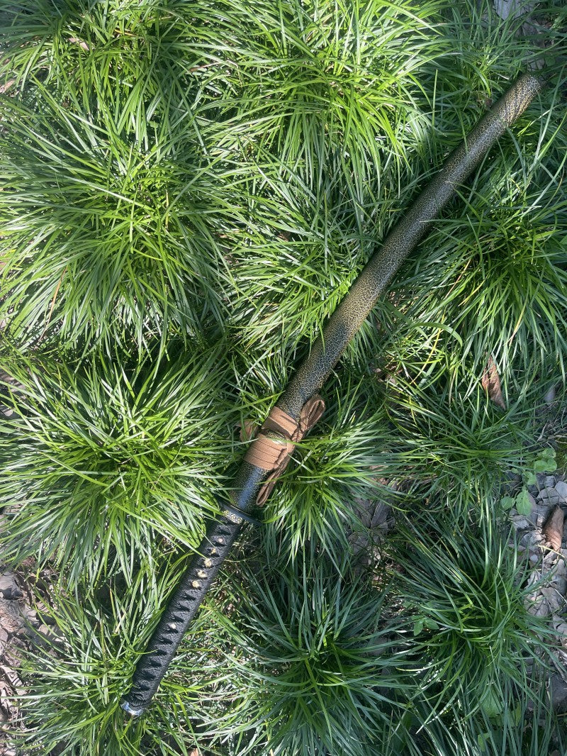 The complete Forest Viper Katana, fully sheathed in its snake-pattern saya, resting in a natural forest setting.